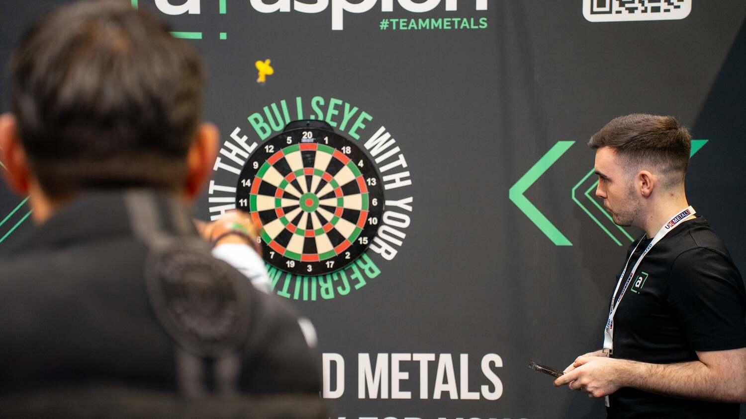 Darts Metals Expo 2025 Aspion Search team member overseeing a darts game at the UK Metals Expo stand, with a branded backdrop reading "Hit the Bullseye with Your Recruitment". Ask ChatGPT